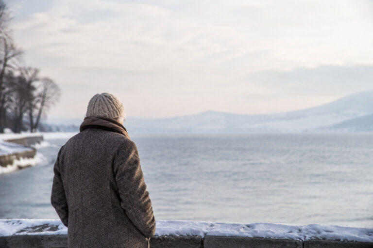 Vue de dos d'une personne âgée avec un manteau et un bonnet, regardant un lac enneigé en hiver.