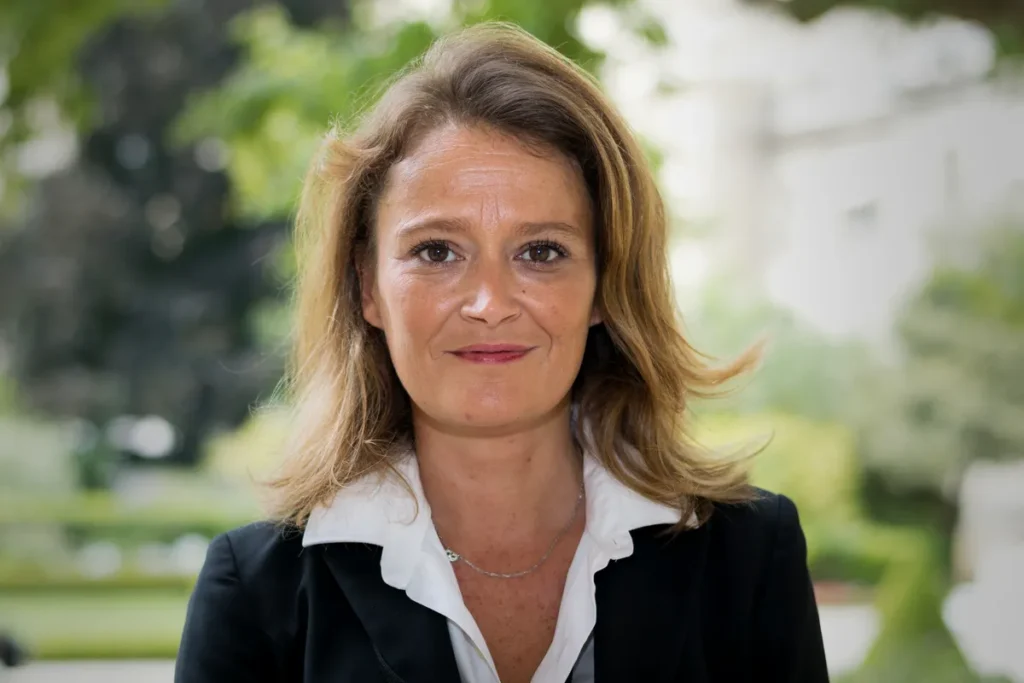 Portrait d’Olivia Grégoire, cheveux châtain clair mi-longs, veste noire sur chemise blanche, souriante face à l’objectif, dans un jardin arboré et flou.